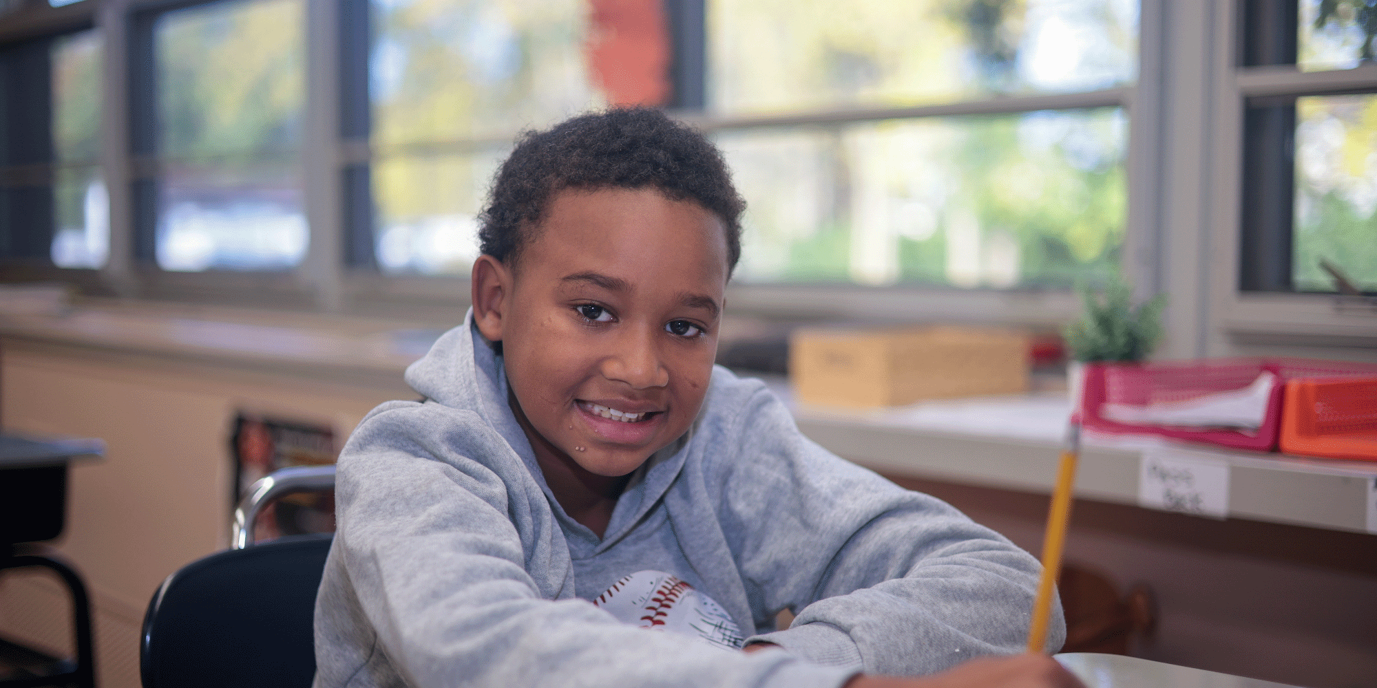 Smiling student at a classroom desk holding a pencil.